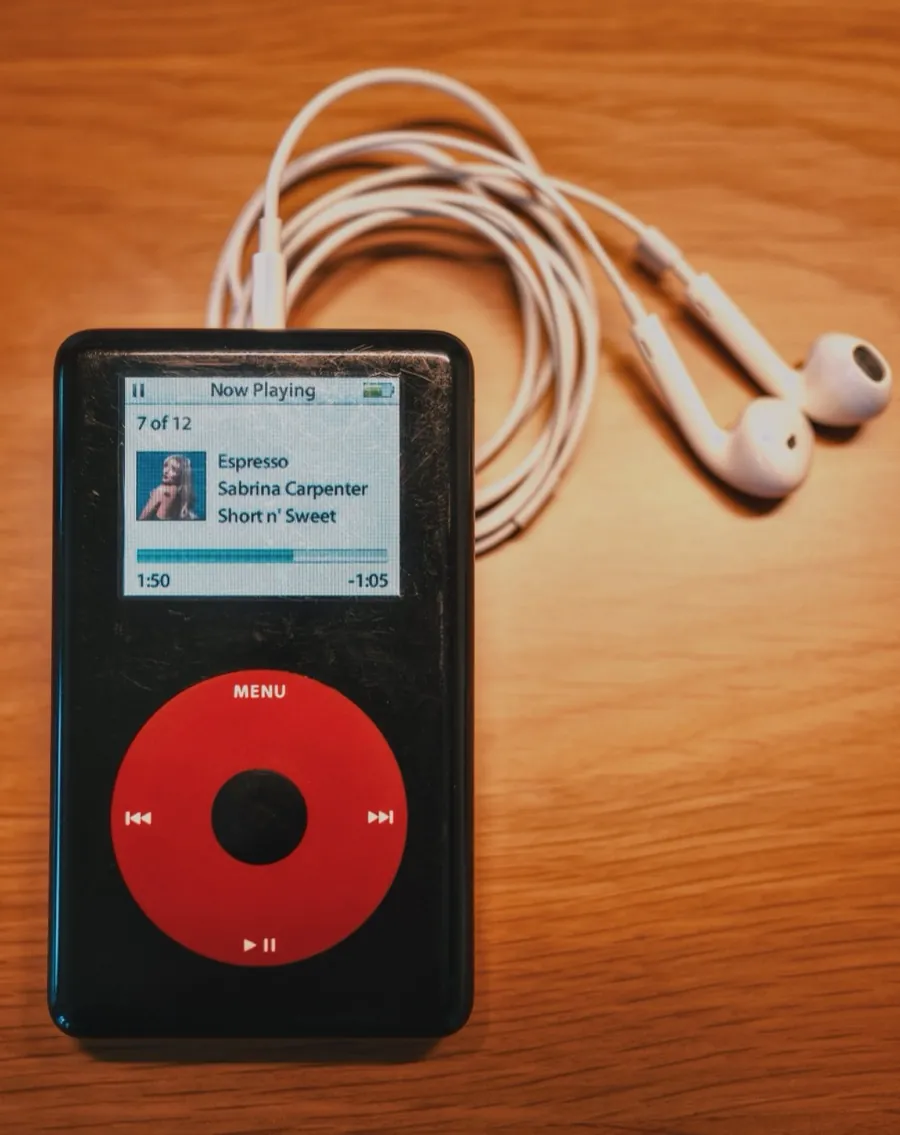 A black iPod with a red click wheel on a wooden desk beside coiled white earphones, its screen showing Now Playing: Espresso by Sabrina Carpenter, from Short n' Sweet, with the album cover displayed.
